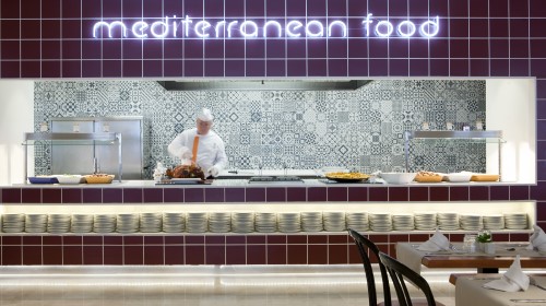 Cocina de comida mediterránea con chef y platos listos para servir. Azulejos decorativos al fondo.